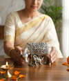 Woman in a white saree holding a decorative metal cow and calf sculpture on a wooden table.